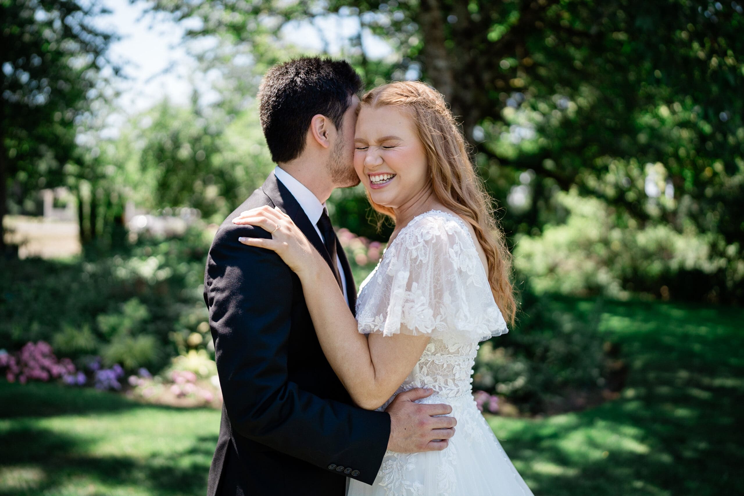 A joyful bride and groom laughing together while a photographer captures the moment from a distance.