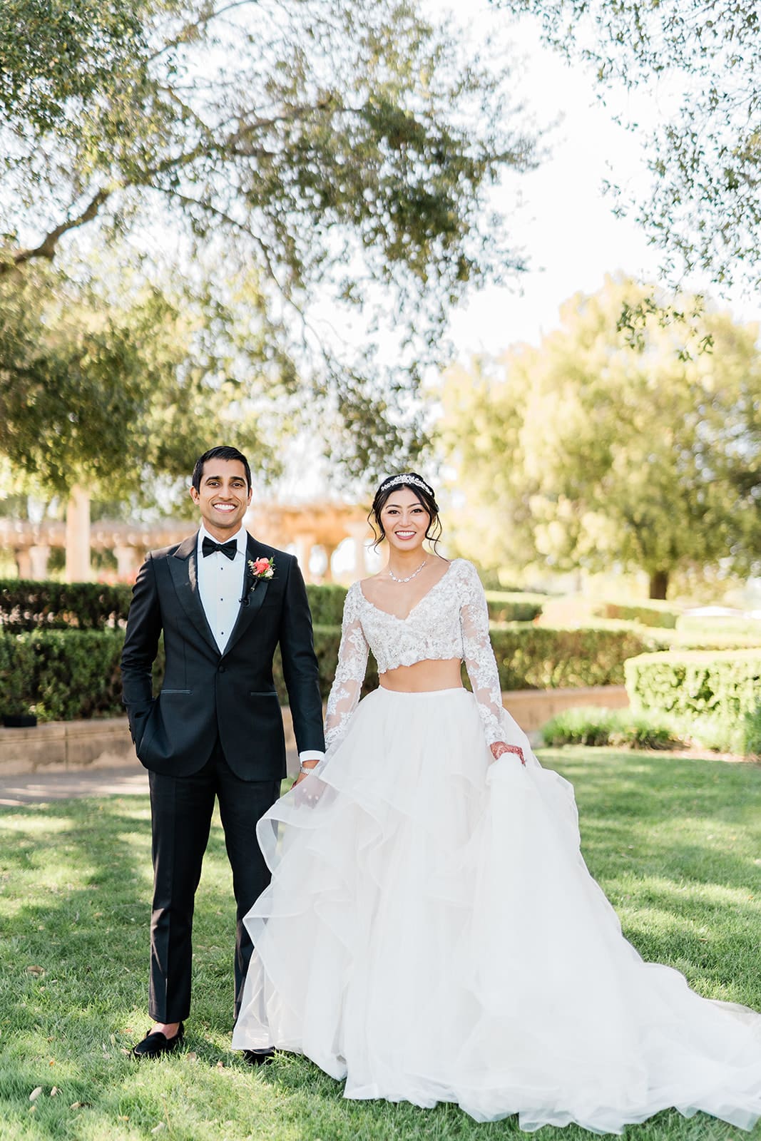 A couple smiling confidently at their photographer with a beautiful outdoor venue in the background.