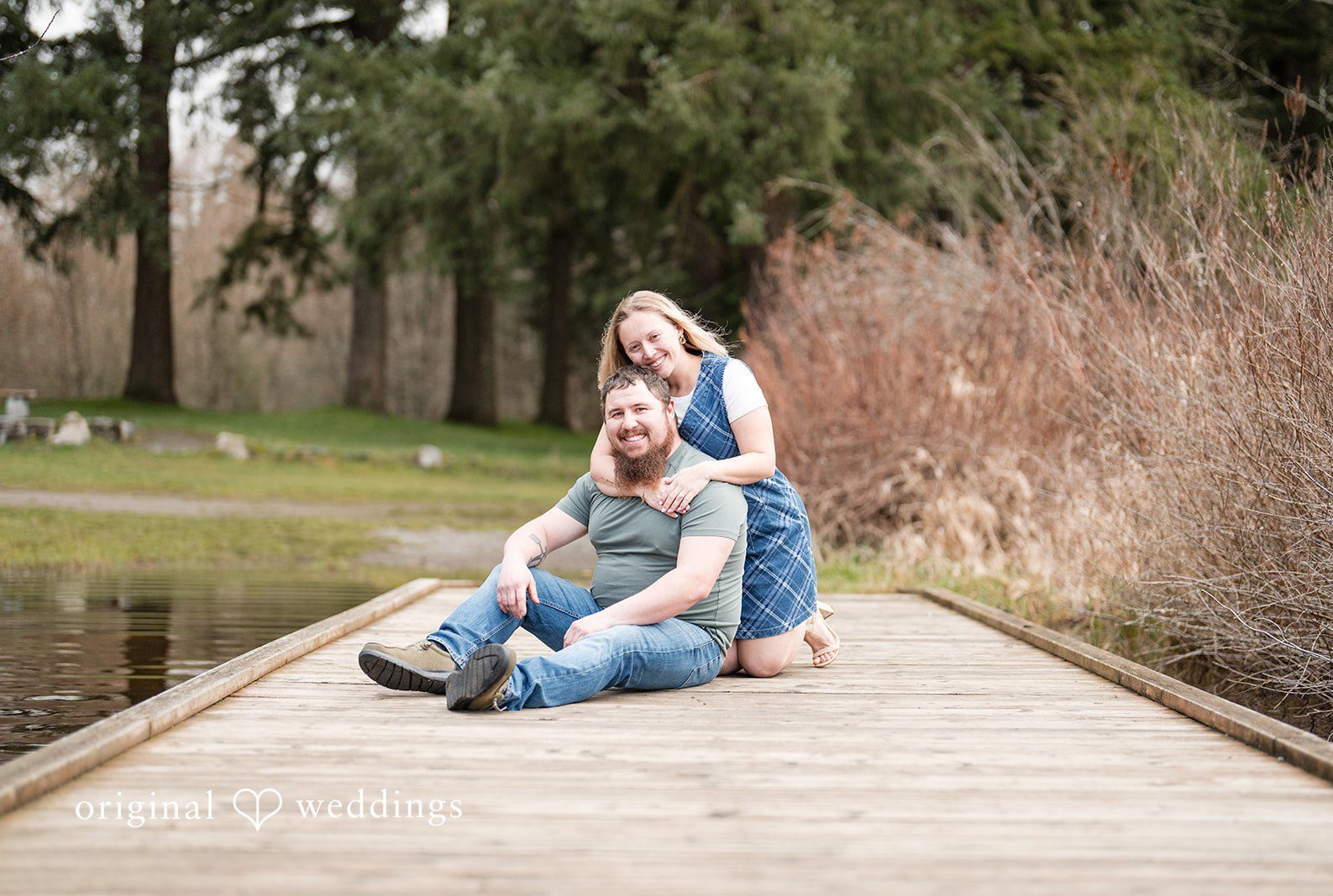 Engagement photography services by Original Weddings capturing bride hugging groom from behind while sitting and smiling at Clark Lake Park