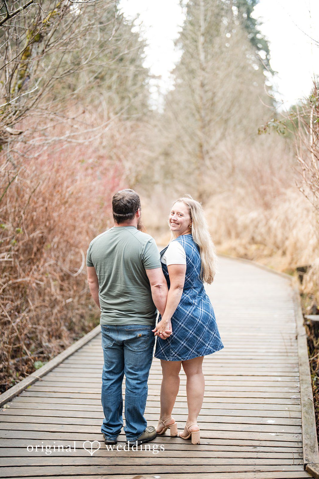 Kayla + Dillon Engagement photography services by Original Weddings capturing couple holding hands with bride looking back at Clark Lake Park