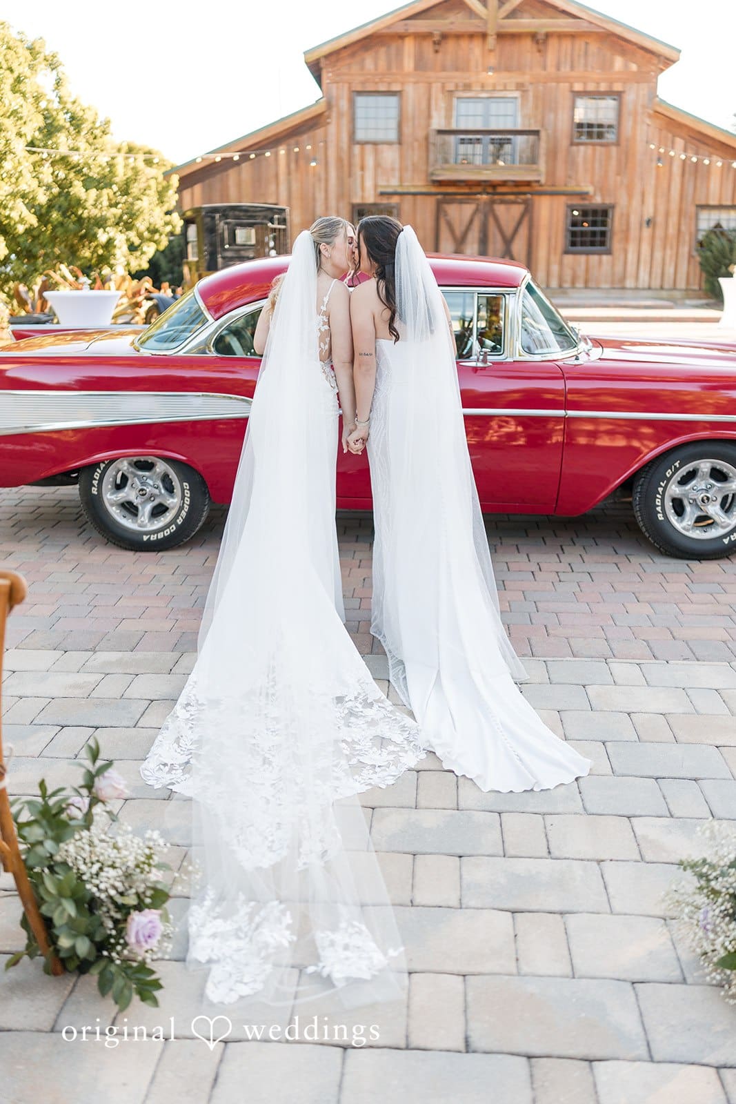 Two brides in white gowns near a red car.