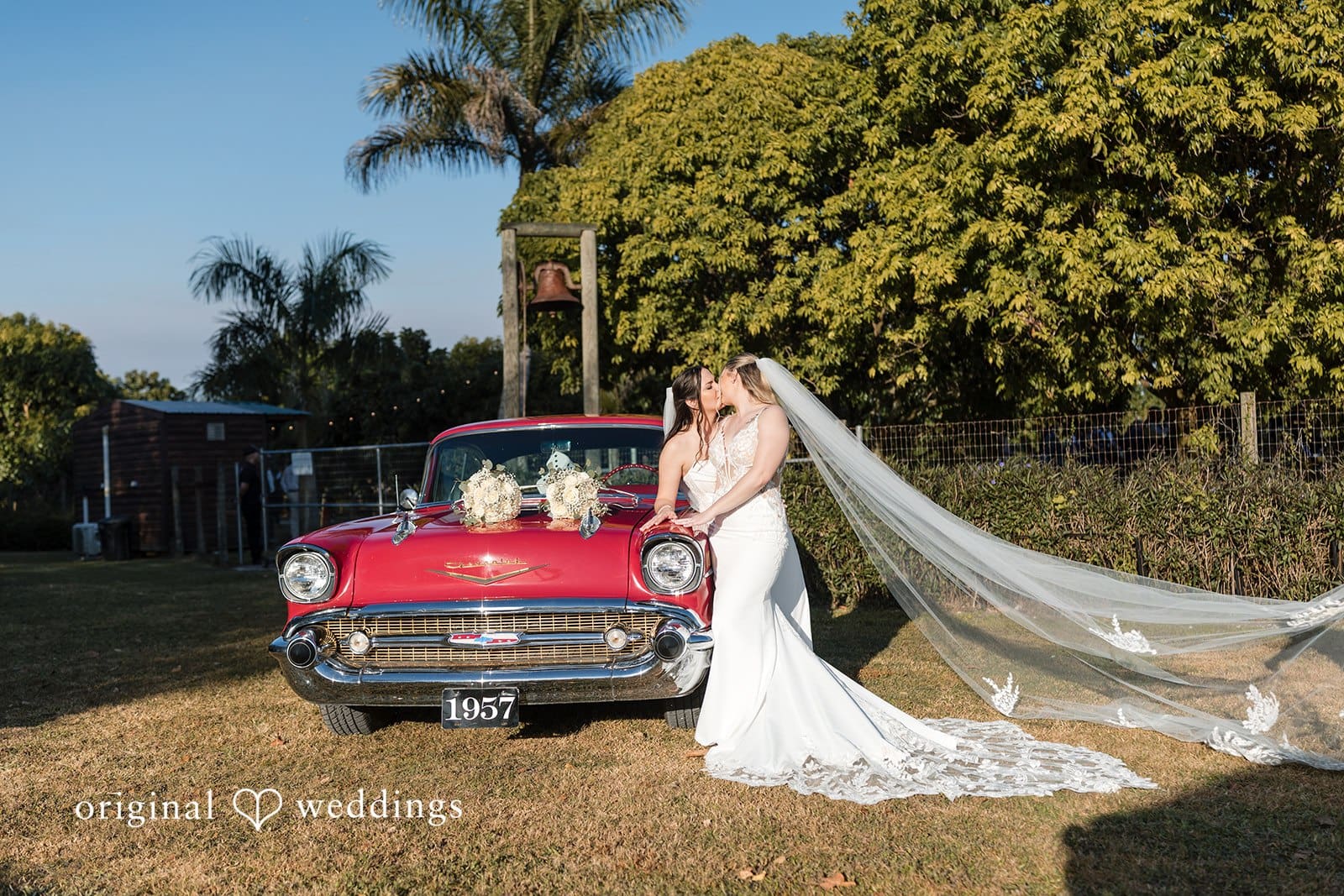 Two brides kissing in front of a red car.