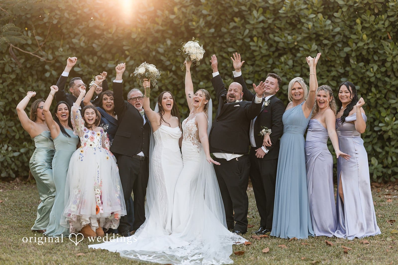 Brides with bridesmaids posing with arms raised in white dresses.
