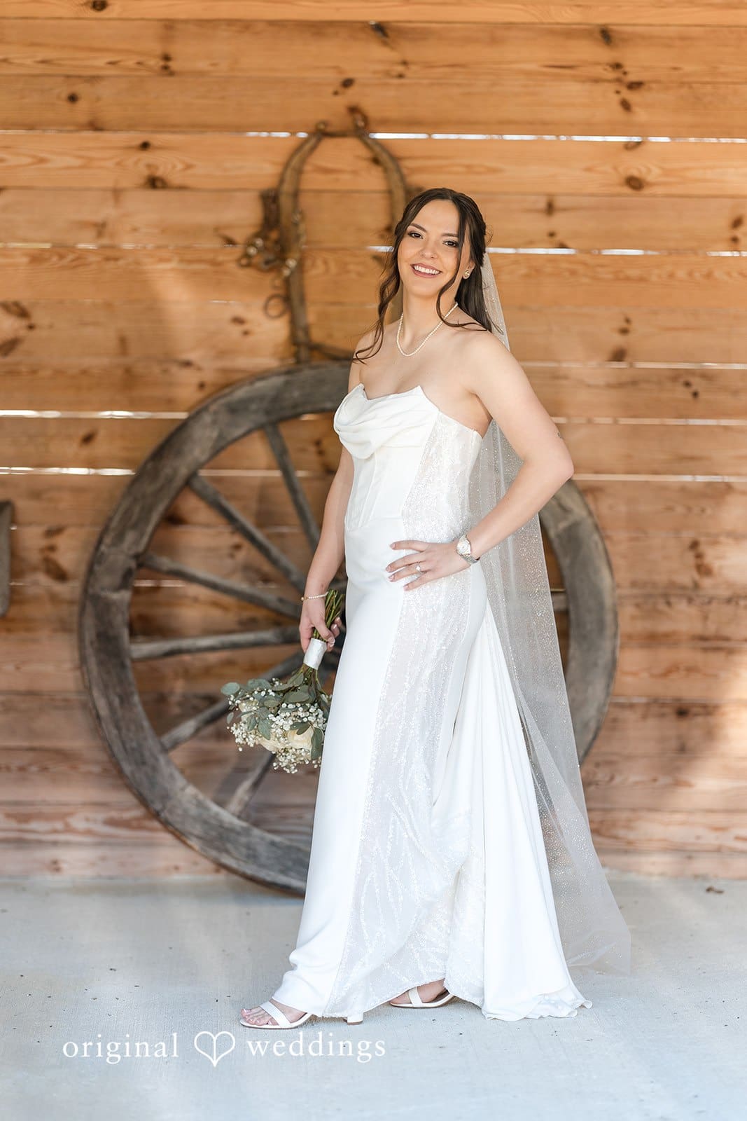 Bride posing by rustic wooden wall with wagon wheel.