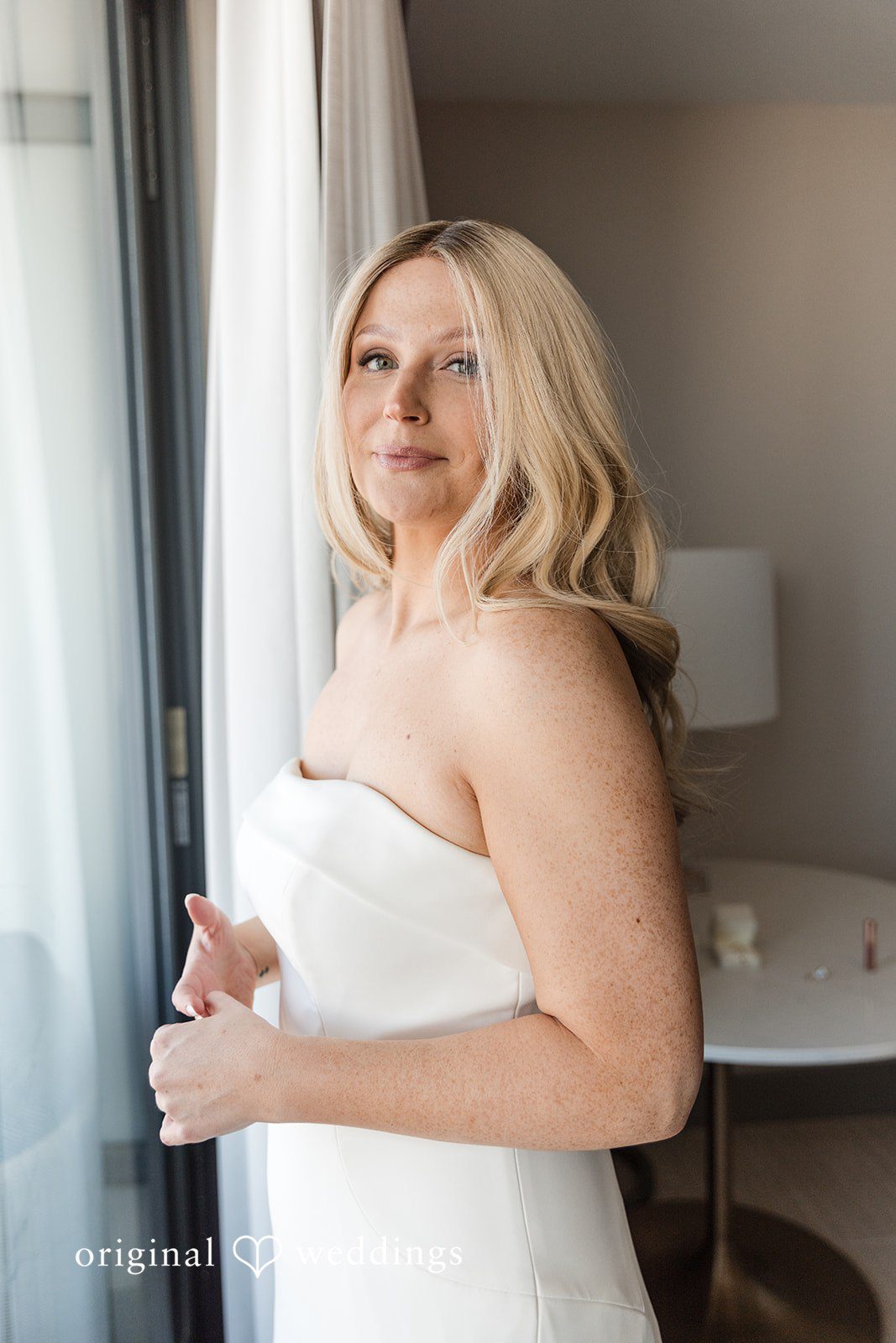 Bride in a white dress standing near a window.