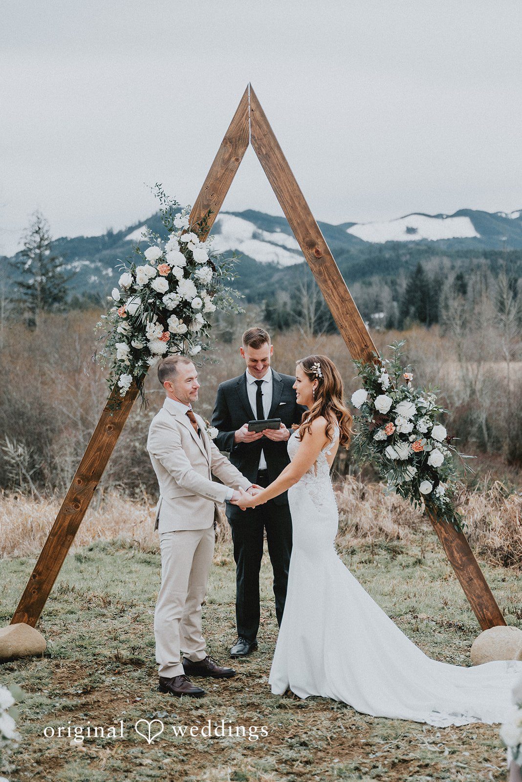 Kelsey + Adam Bride & groom vowing under a floral triangular arch with mountains behind.