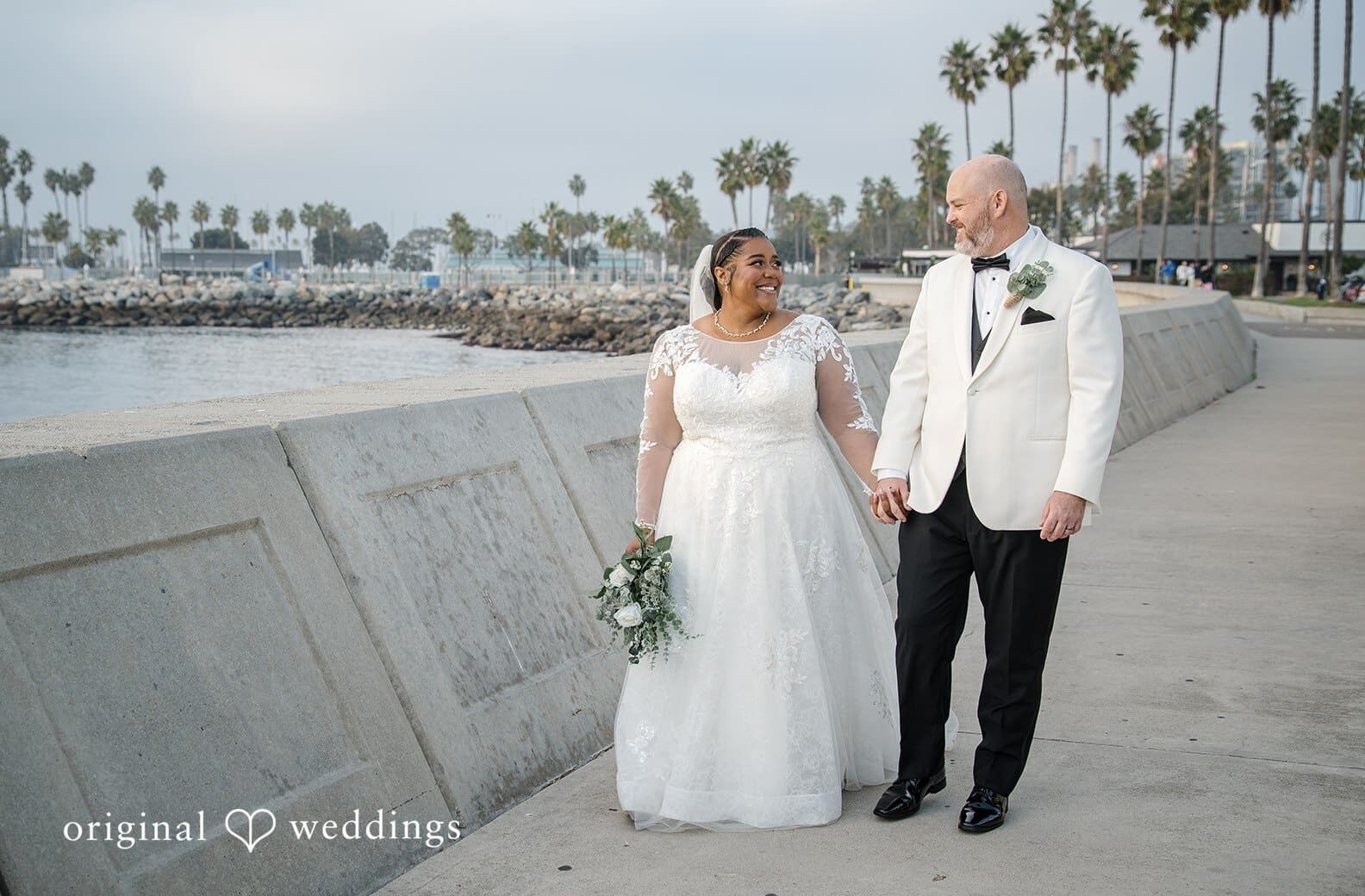 A couple walking hand in hand along a coastal walkway with palm trees and ocean views, captured by Los Angeles wedding photographers at a beachfront wedding venue.
