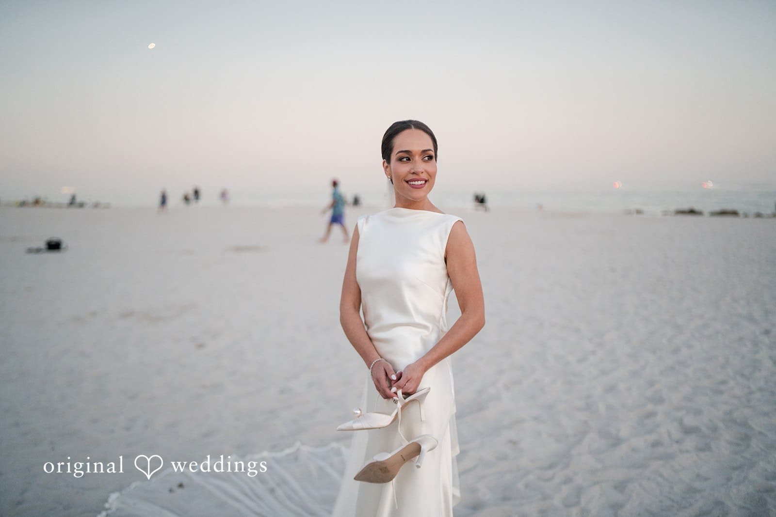 Bride near the ocean holding her shoes on wedding day