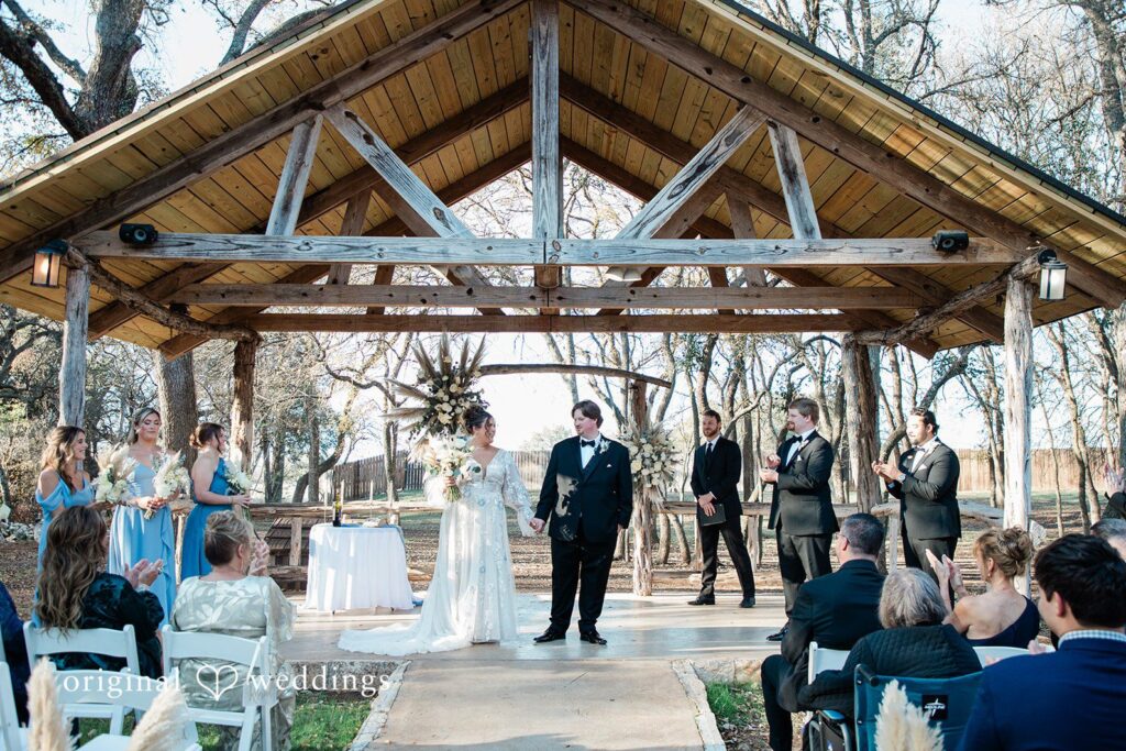 The Texas Old Town Wedding | Kelsey & Riley 1 Under a rustic wooden pavilion, the couple stands hand in hand as guests applaud during their ceremony, beautifully captured through Austin wedding photography.