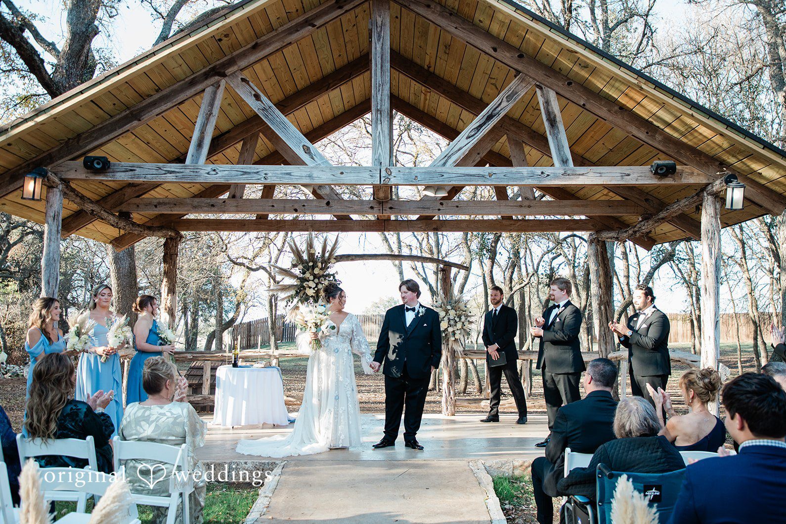 Under a rustic wooden pavilion, the couple stands hand in hand as guests applaud during their ceremony, beautifully captured through Austin wedding photography.