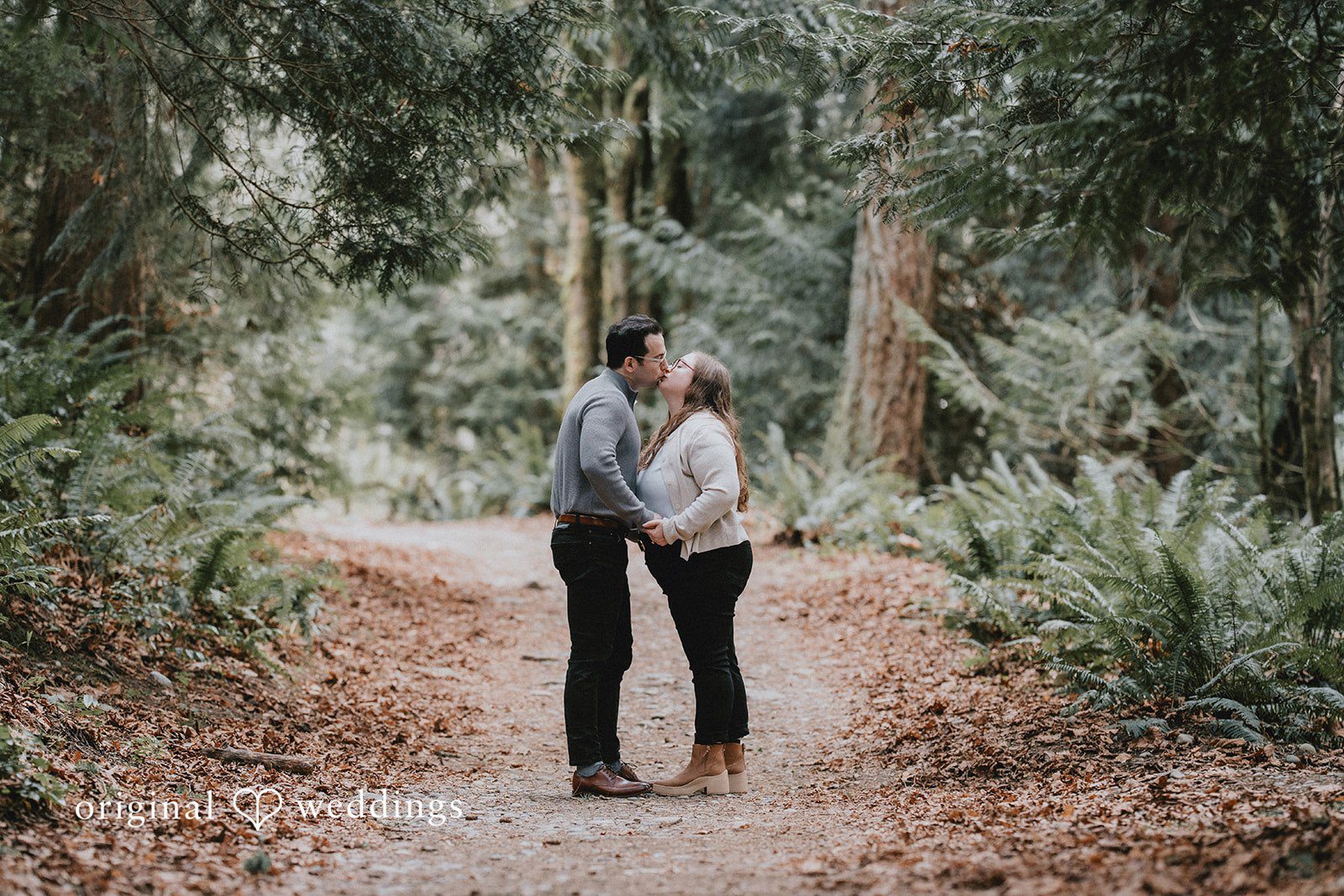 Couple kissing on a forest path, surrounded by trees and greenery.