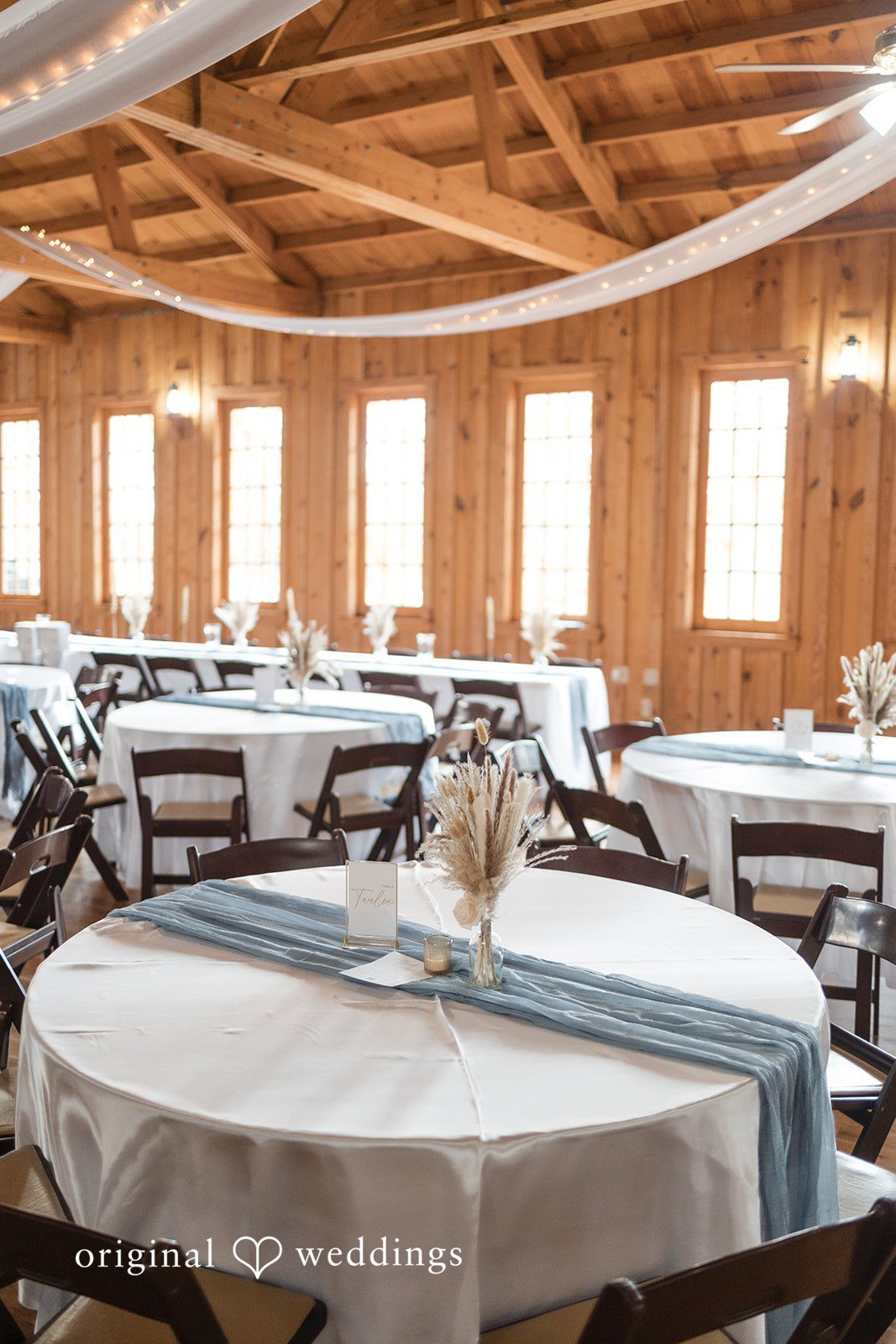 Kelsey + Riley Round tables decorated with simple centerpieces fill a bright wooden venue.