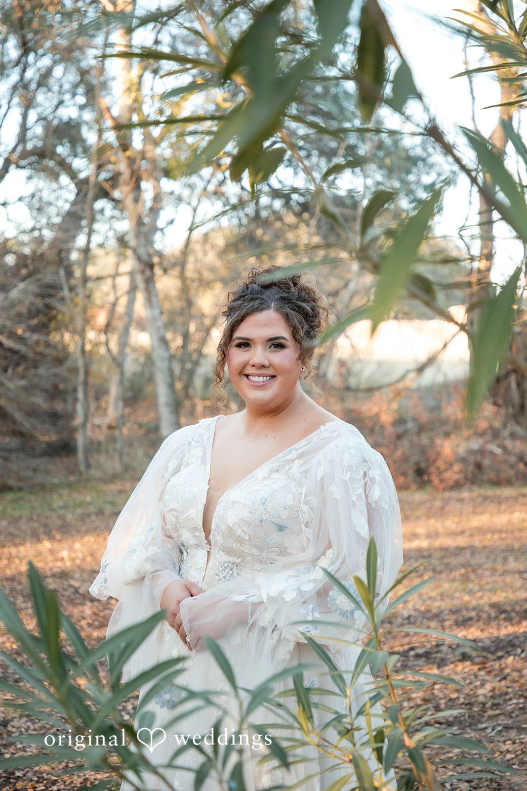 Kelsey + Riley The bride smiles while standing among greenery in her wedding dress.