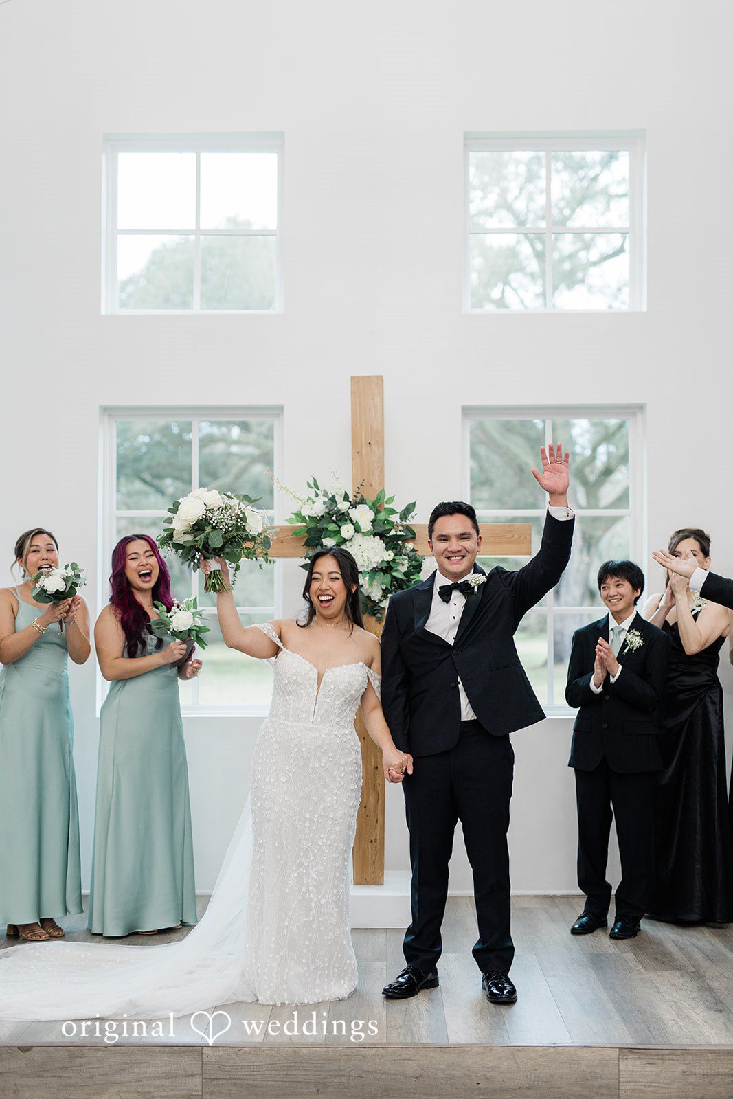 Dominic + Erika Joy Couple in wedding attire raising their hands in joy at the altar as their wedding party claps.