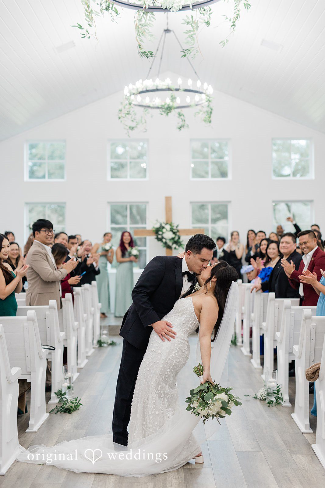 Dominic + Erika Joy Couple sharing a kiss in the center of an aisle lined with white chairs and seated guests.