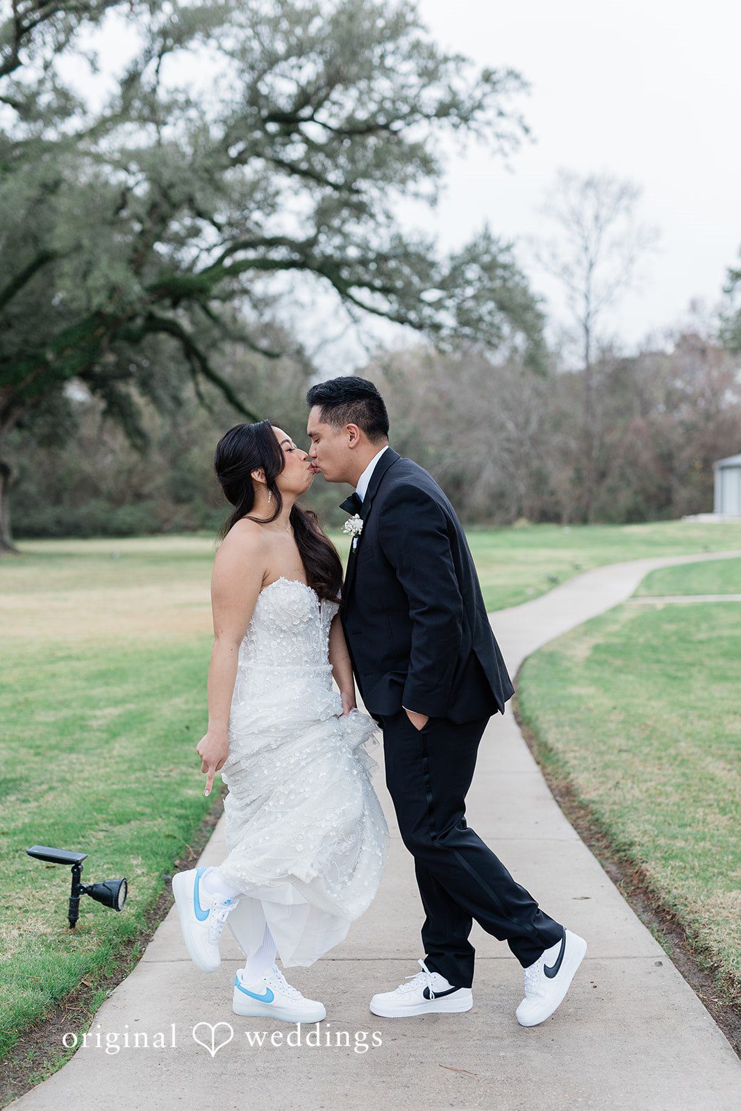 Dominic + Erika Joy Couple in wedding attire and sneakers kissing on a paved path.