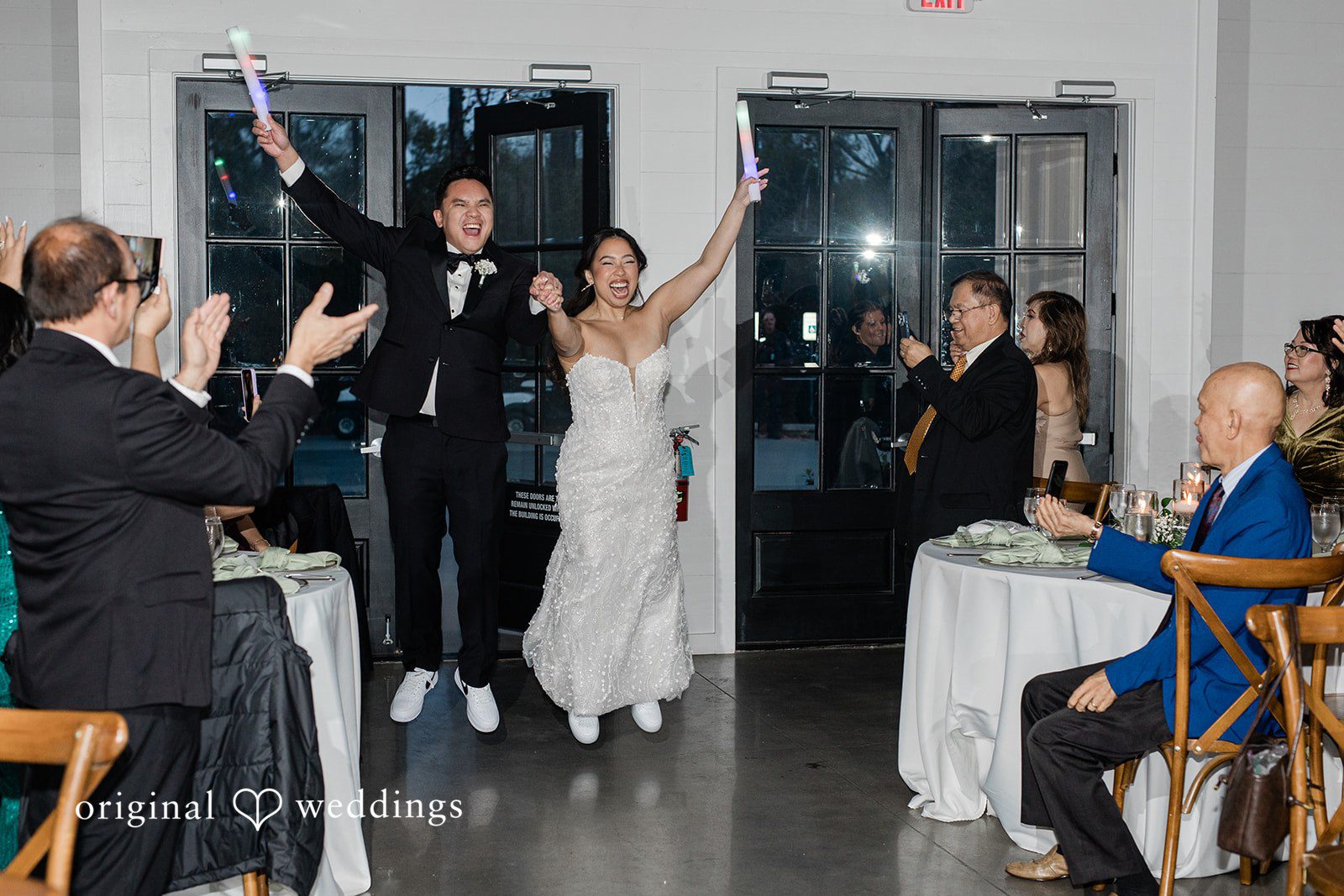 Dominic + Erika Joy Couple in wedding attire entering a reception room with arms raised while guests clap