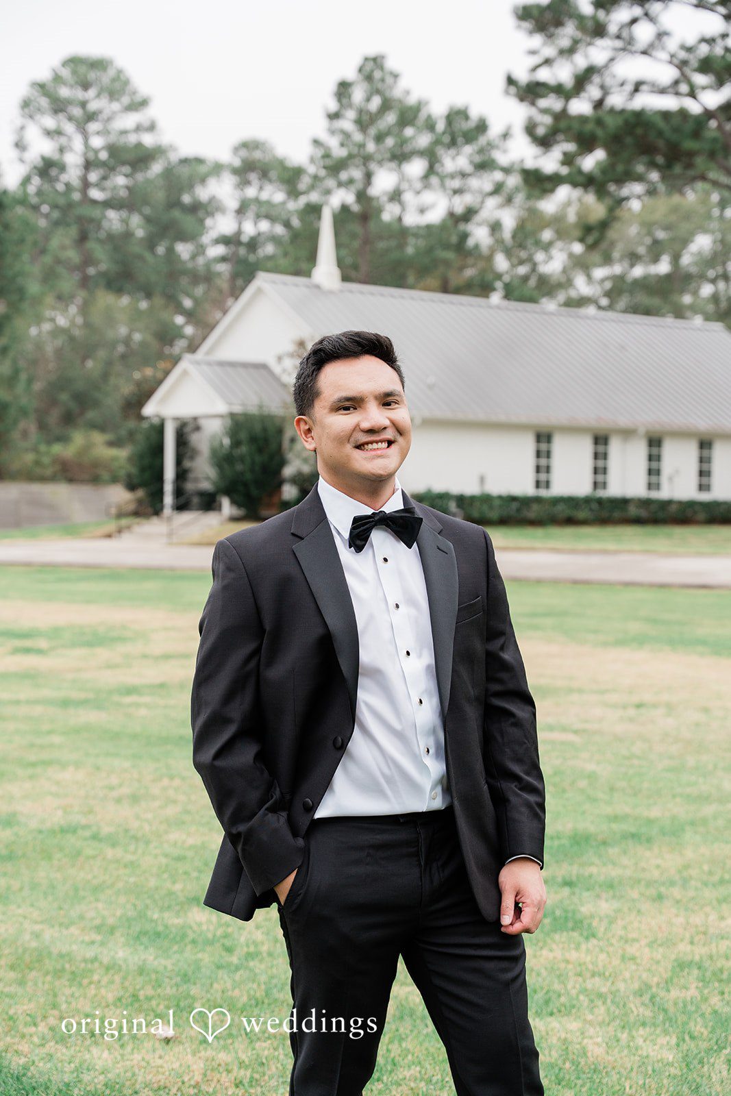Dominic + Erika Joy Groom dressed in a classic black tuxedo with a black bowtie, standing on a manicured lawn.