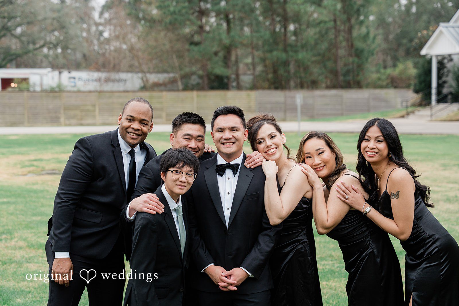 Dominic + Erika Joy A group of men and women in formal black attire smiling together for a group photo.