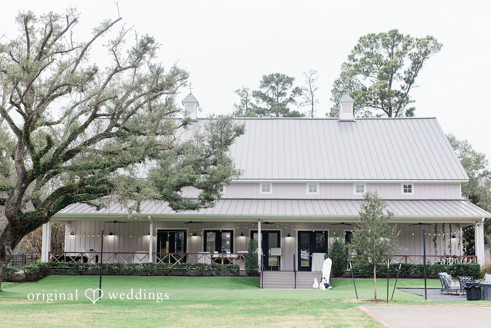 Dominic + Erika Joy A large, rustic white barn-style building with multiple gables, set against a backdrop of tall, green trees.