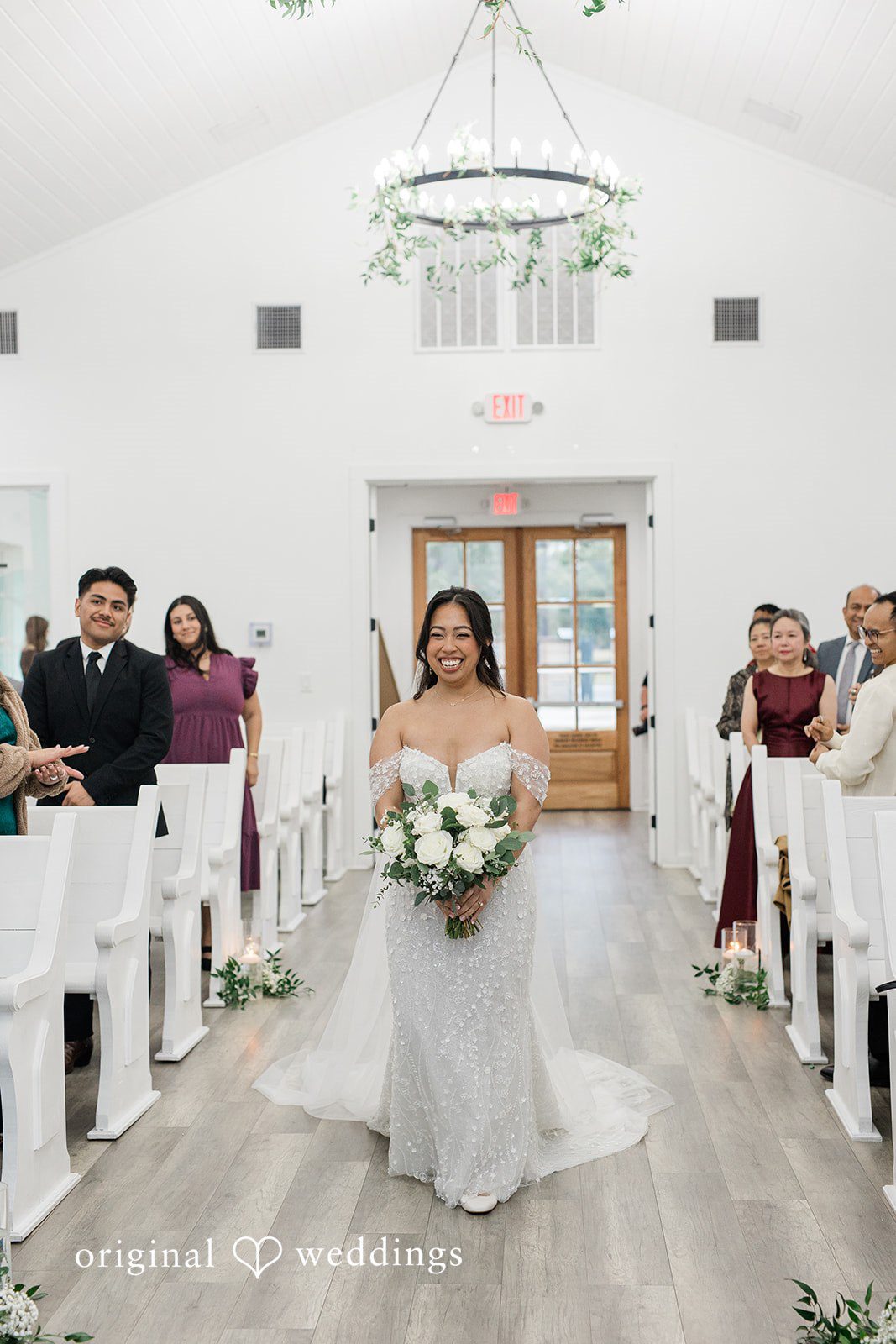 Dominic + Erika Joy A bride in a white gown walking down a church aisle lined with white wooden pews and floral arrangements.