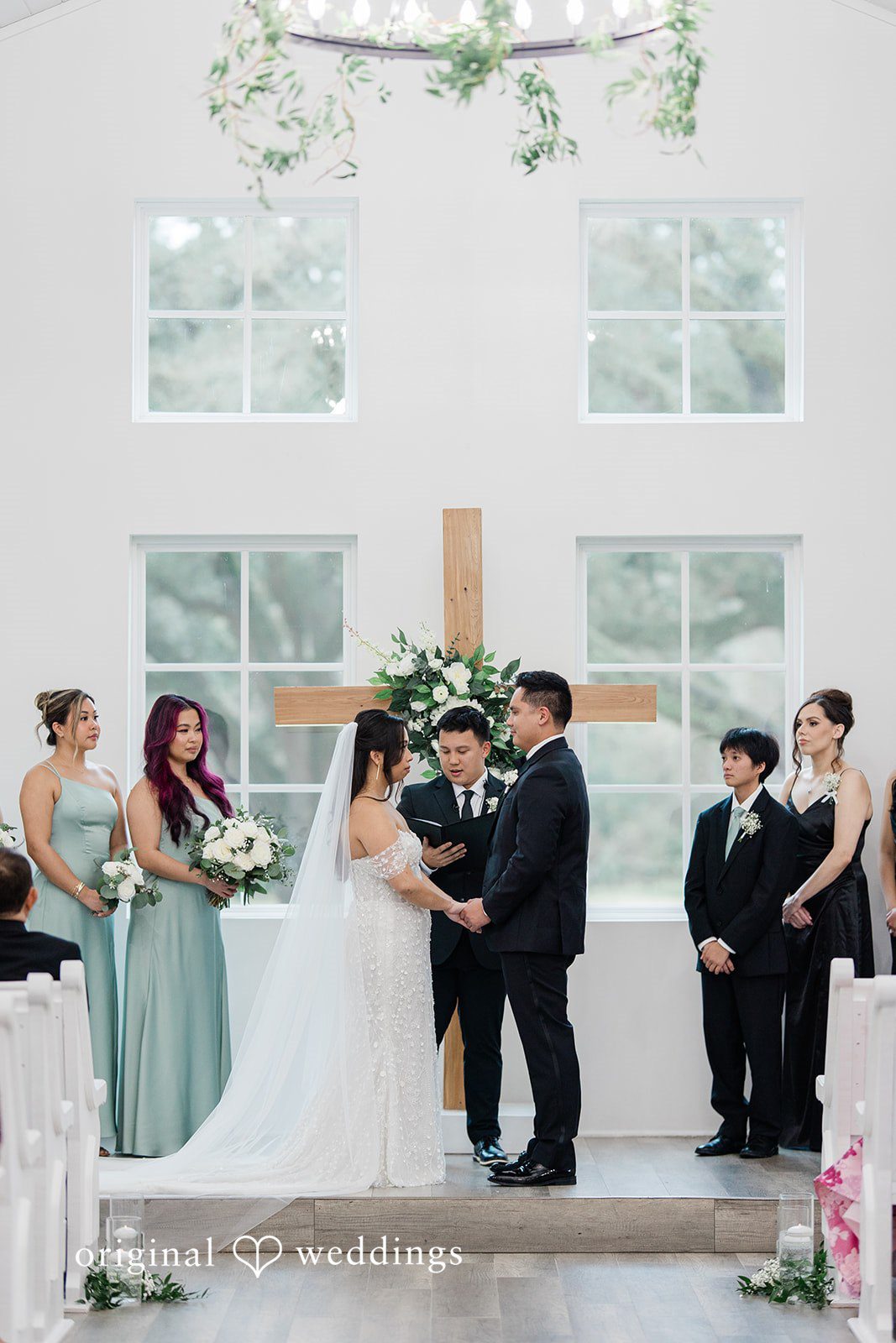 Dominic + Erika Joy Two people standing at an altar under a wooden cross, surrounded by members of the wedding party.