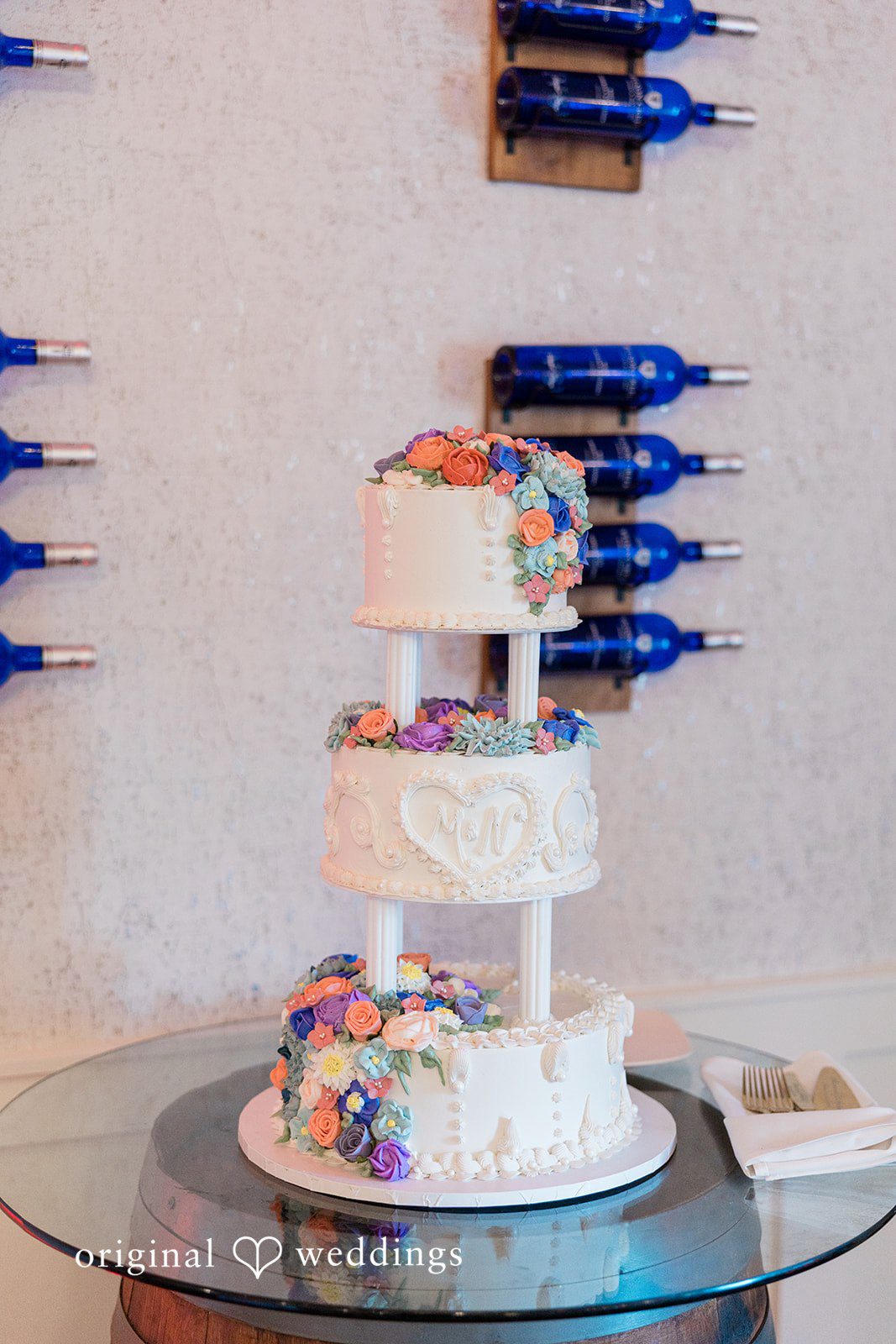 Three-tiered wedding cake decorated with colorful flowers, displayed on a glass table.