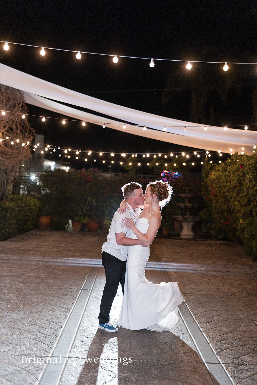 Bride and groom kissing at night under rows of hanging string lights and draped fabric.