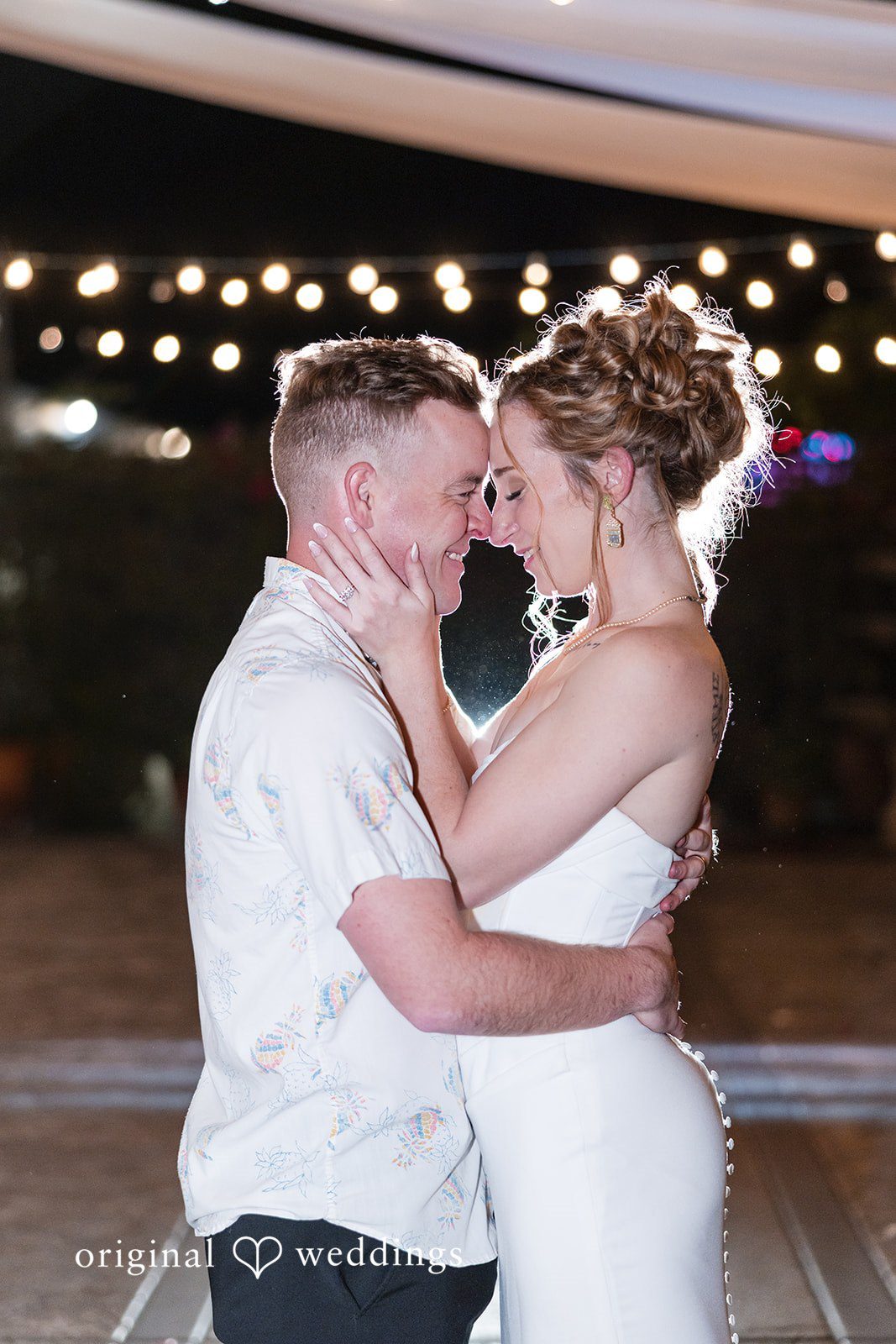 Close-up of a smiling bride and groom embracing face-to-face with blurry string lights in the background.