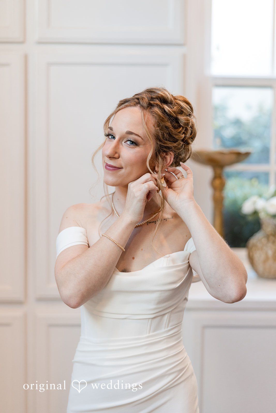 Bride wearing an off-the-shoulder gown while adjusting her earring.