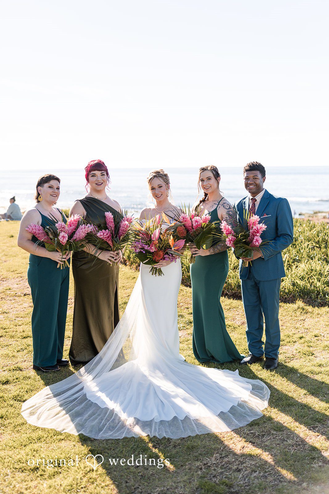 Bride posing with bridesmaids in matching dark green dresses on a grassy field near the ocean.