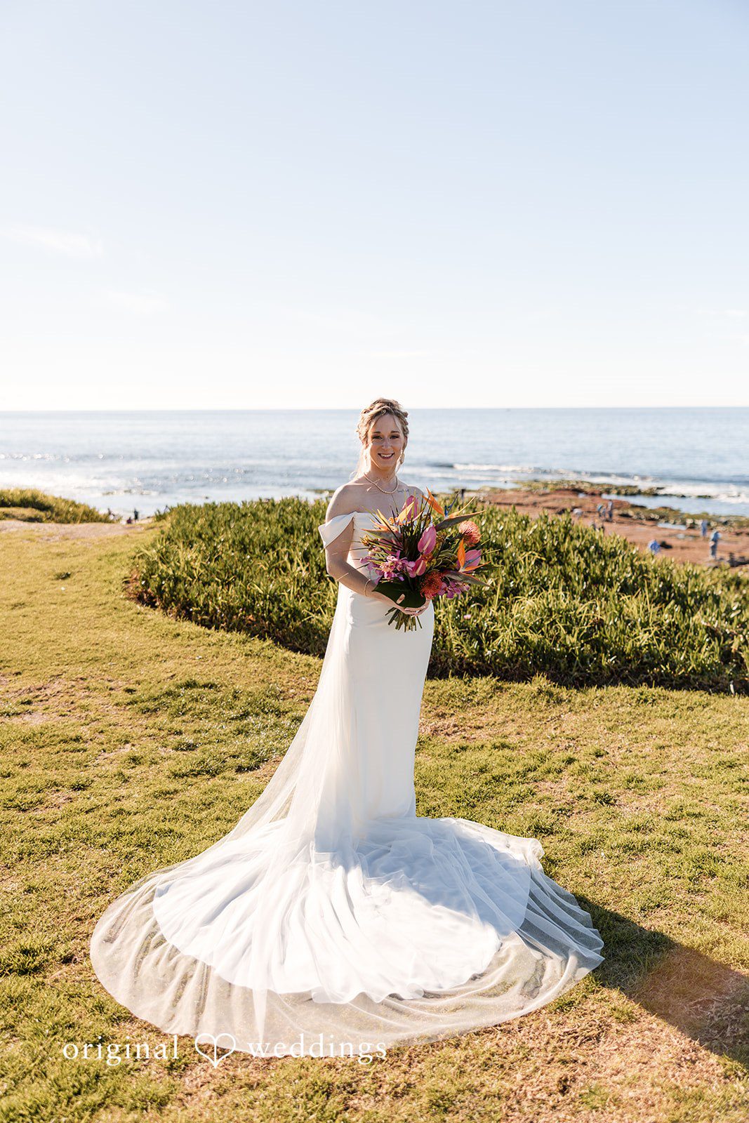 Bride standing on a grassy coastal cliff with a long train spread across the grass.