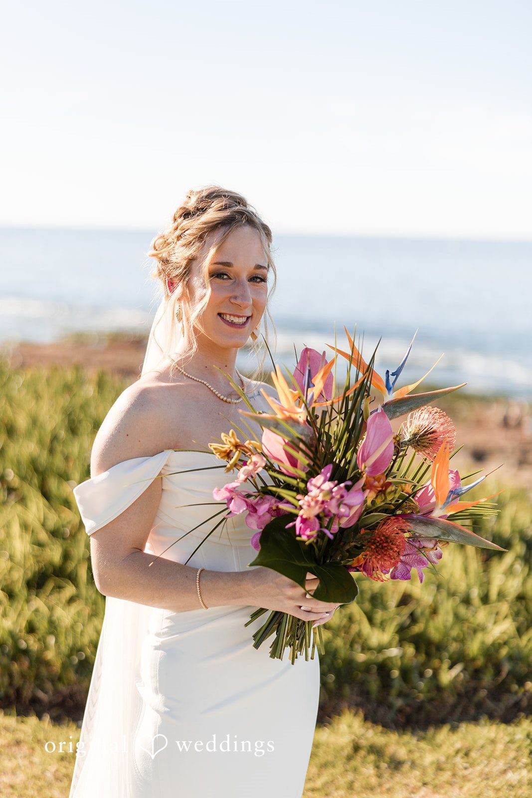 Close-up of a smiling bride holding a lush bouquet of pink tropical flowers.