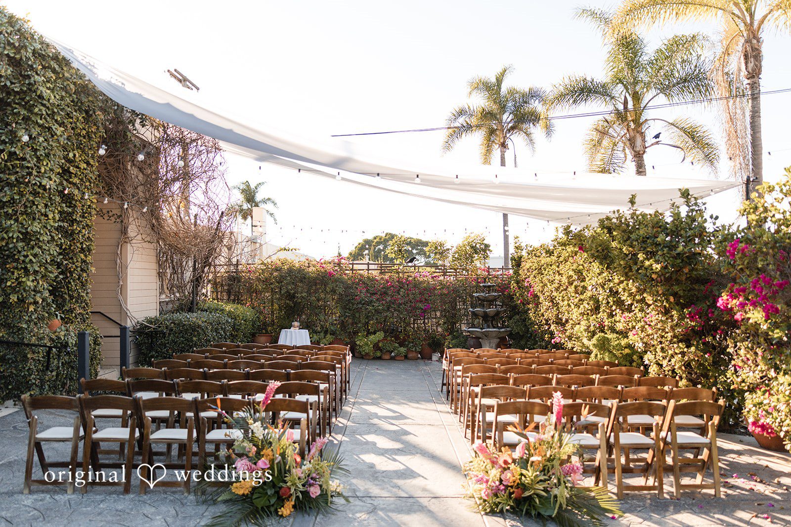 Wedding ceremony aisle flanked by rows of wooden chairs under palm trees.