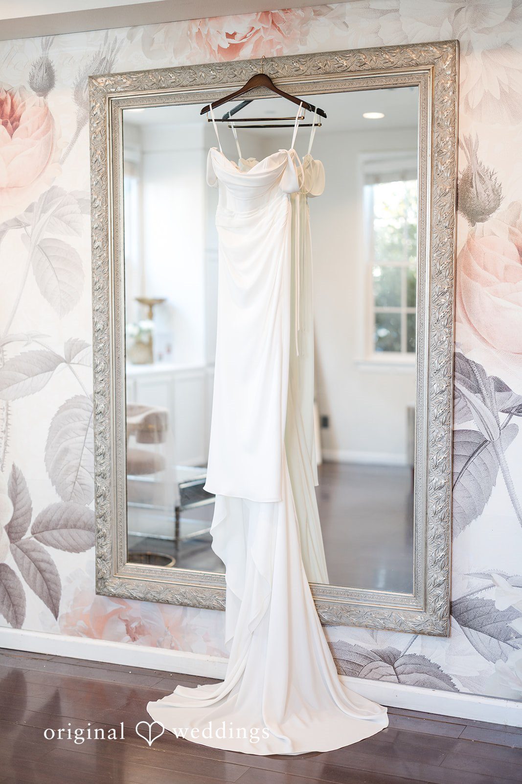 White wedding gown hanging vertically in front of a large framed mirror.