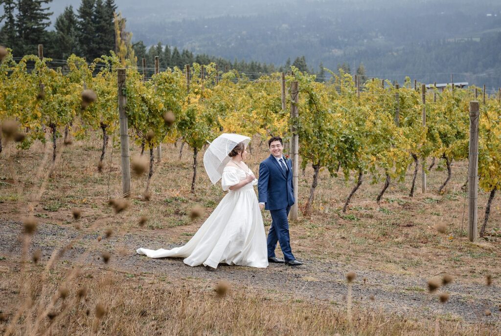 Tiare + Jeremy Bride and groom walking through a vineyard while holding a clear umbrella during their rainy wedding day at Gorge Crest Vineyards captured by Portland wedding photographers from Original Weddings
