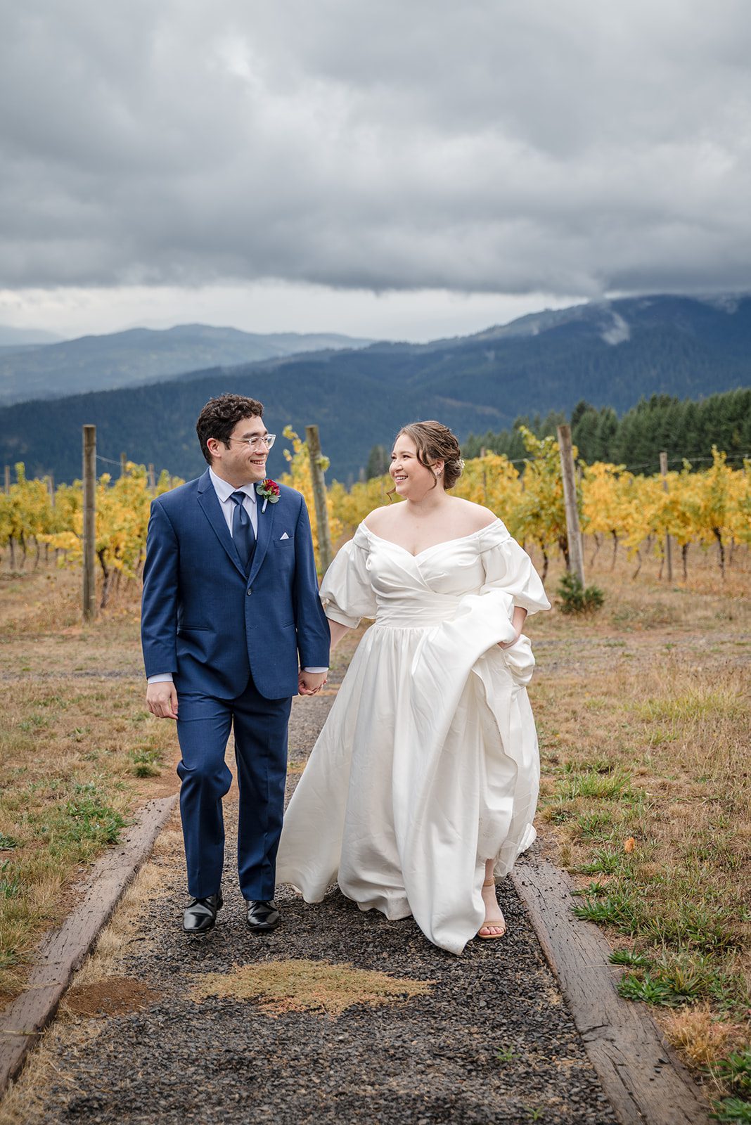 Tiare + Jeremy Bride and groom walking hand in hand along a vineyard path with mountains in the distance