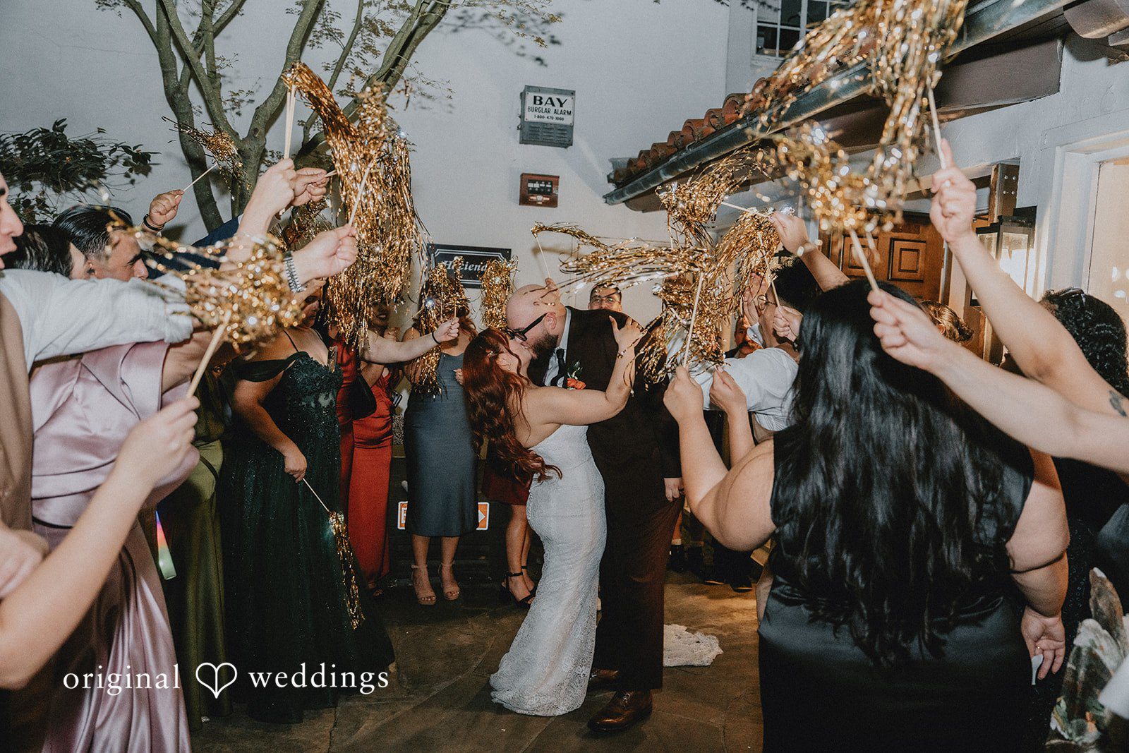 Bride and groom share a kiss under gold streamers while guests surround them