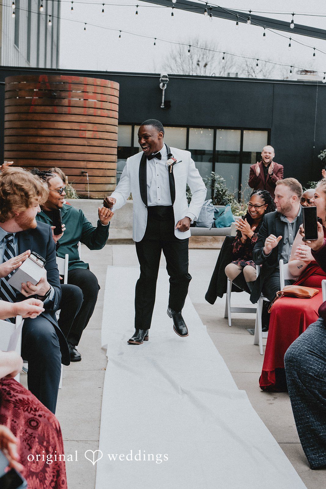 A groom in a white tuxedo jacket walks down a white aisle runner between seated guests.
