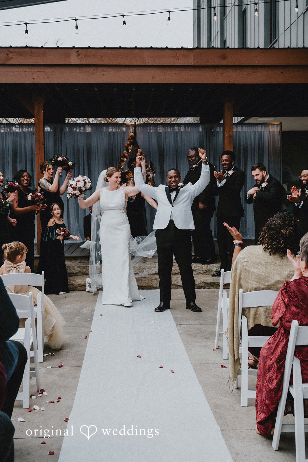 The newlyweds walking down the white aisle runner, cheering with their hands raised.