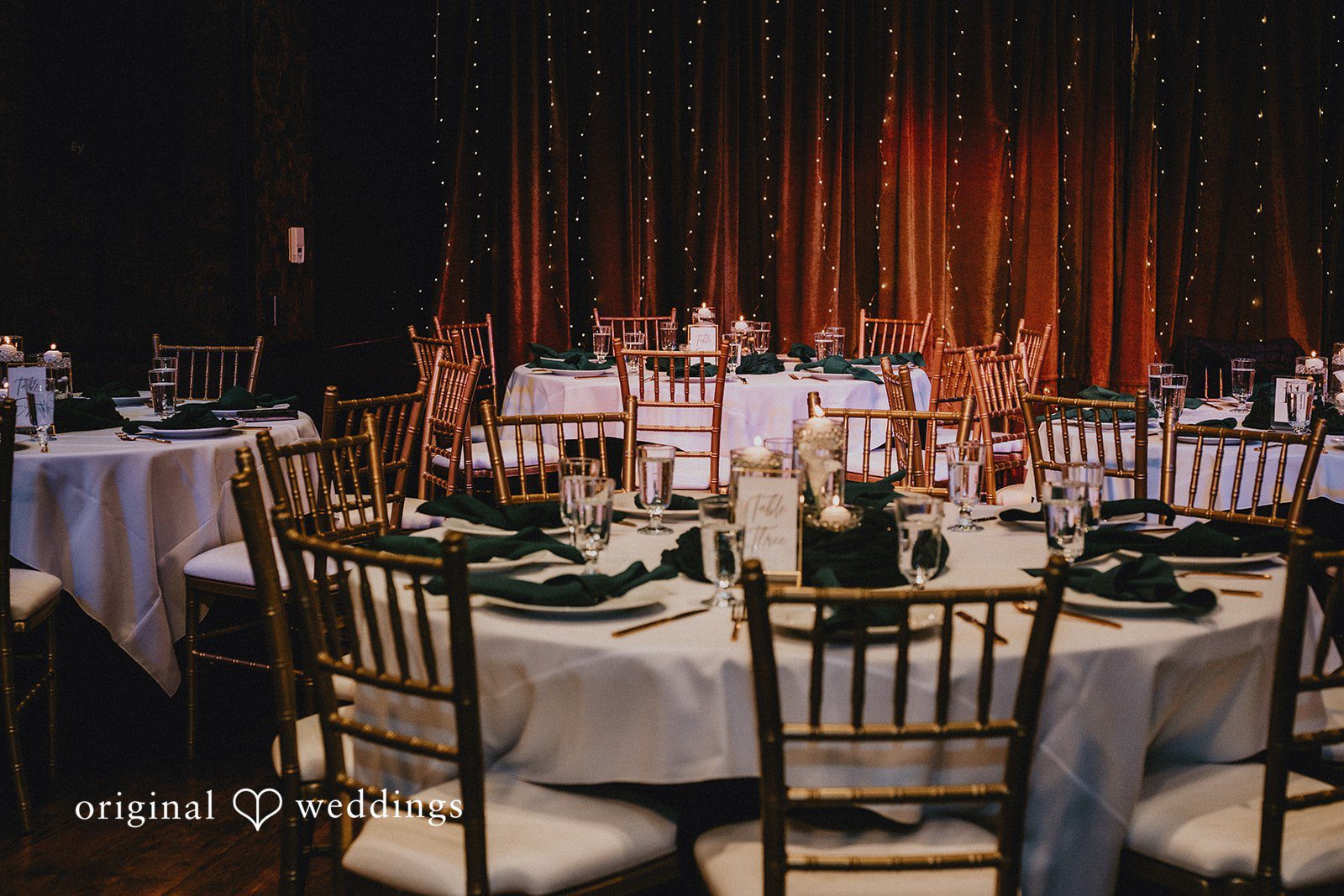 Multiple round banquet tables with white cloths and gold chairs in a dimly lit reception hall.
