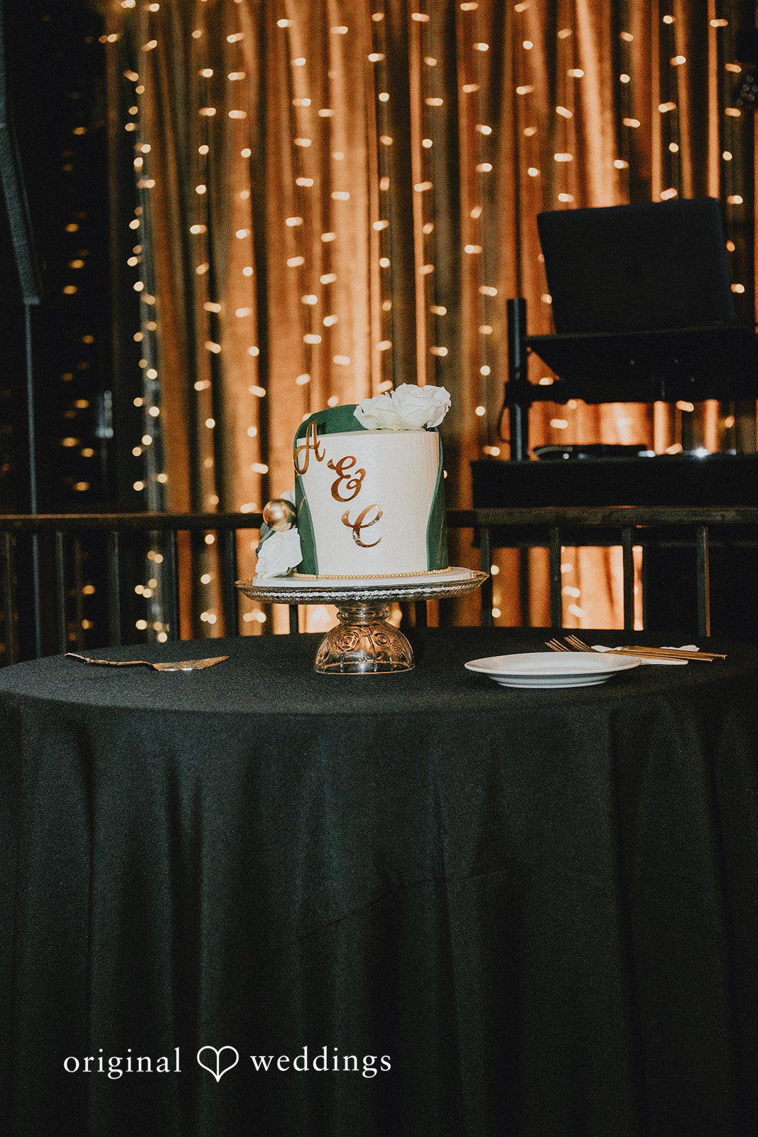 A small, tiered wedding cake with floral accents sits on a dark-clothed table.