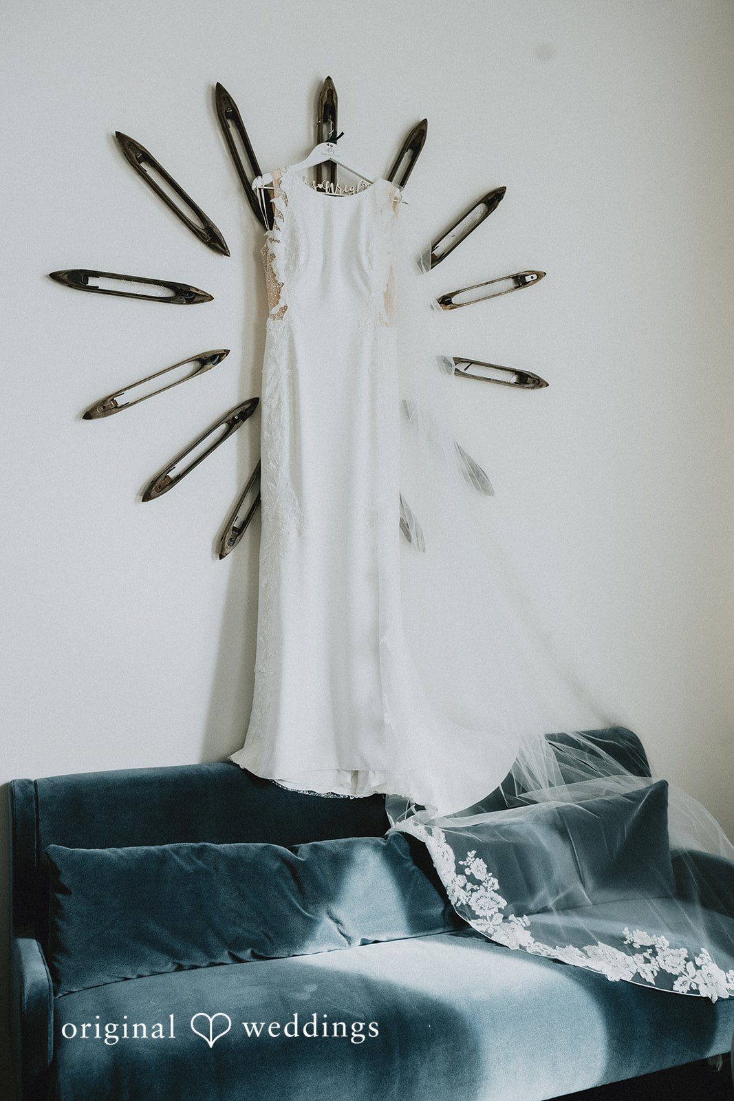 A white wedding dress hangs against a wall decorated with a circular pattern of metallic objects.