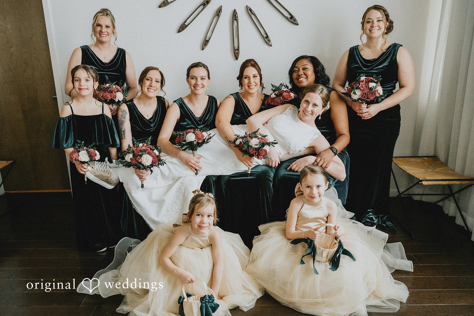 A group of bridesmaids in dark dresses and flower girls in white gowns posing together.