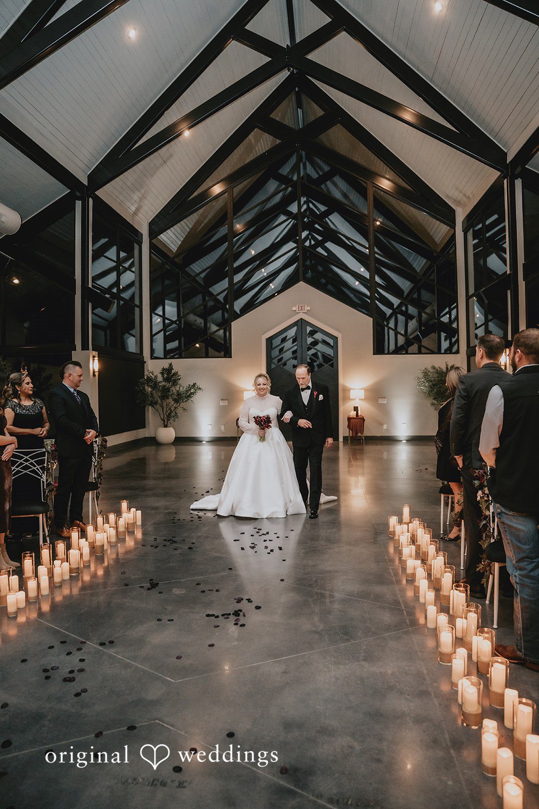 Bride walking down a wide aisle lined with glowing candles toward the altar in a modern chapel.