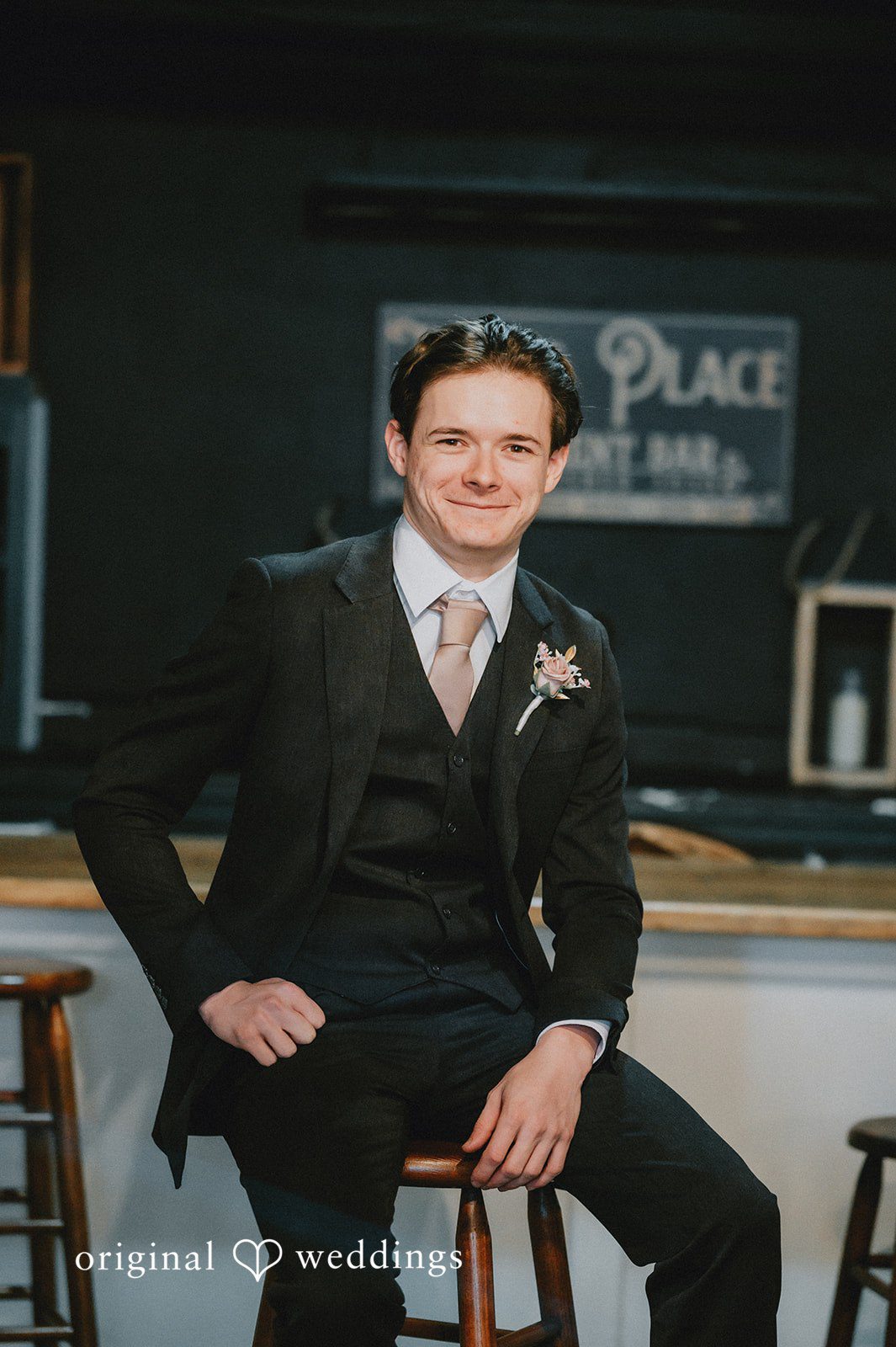 Ayana + Gabriel A groom smiling while seated in his wedding suit against a dark background.