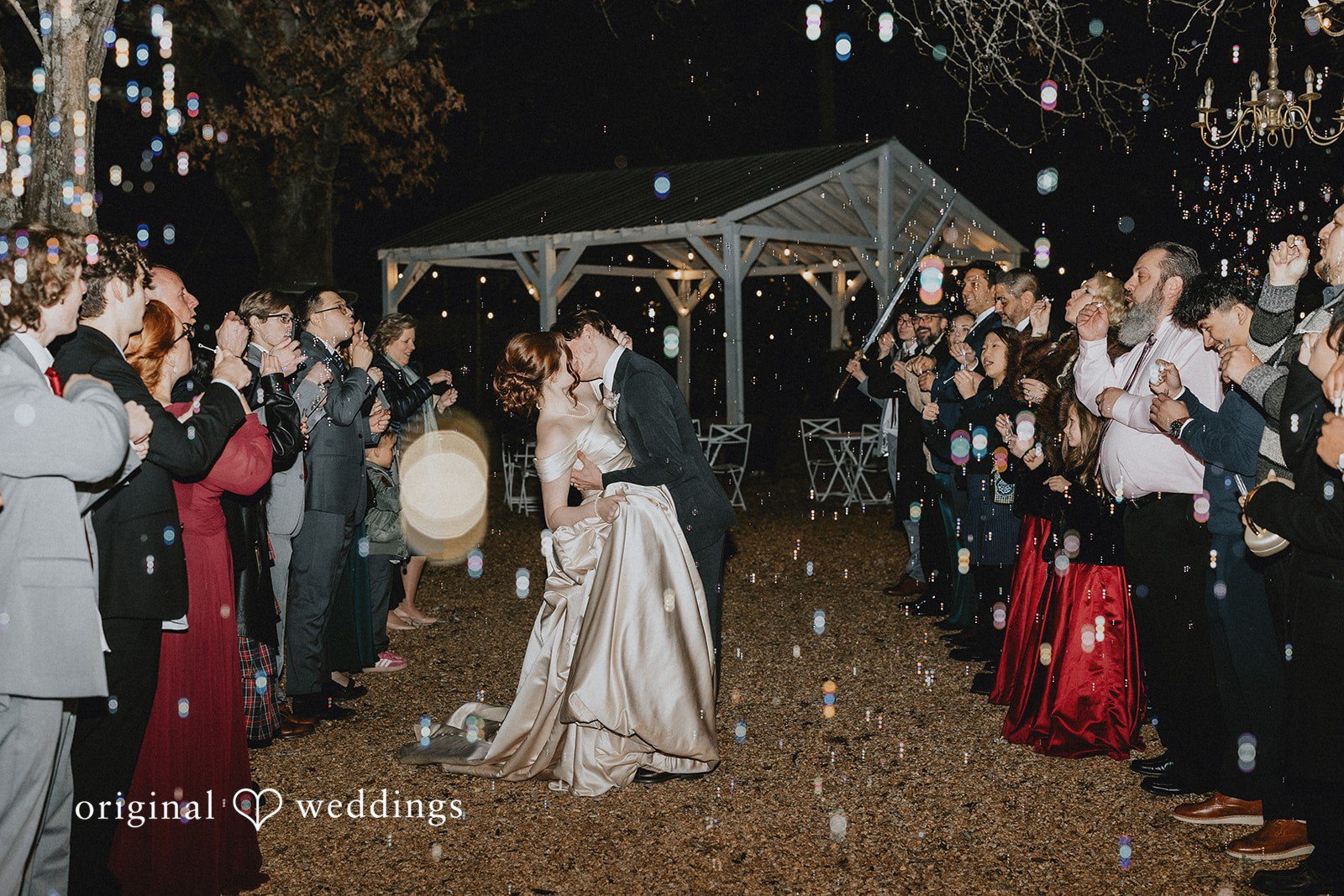 Ayana + Gabriel Bride and groom sharing a dipping kiss during a nighttime bubble send-off surrounded by wedding guests.