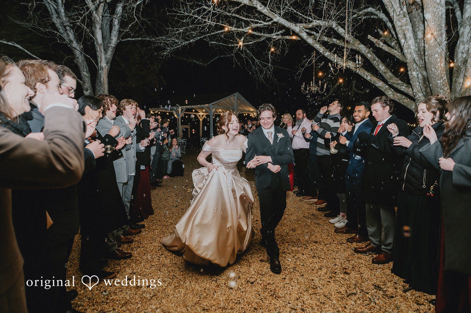Ayana + Gabriel Couple walking hand-in-hand while laughing during a nighttime bubble grand exit surrounded by cheering wedding guests.