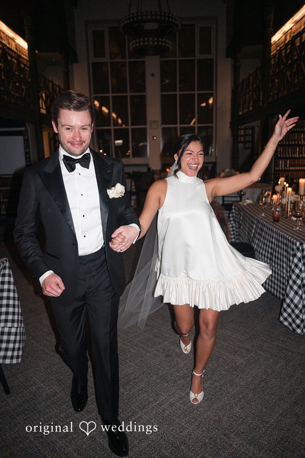 A groom in a tuxedo and a bride in a short dress dancing together into the reception room.