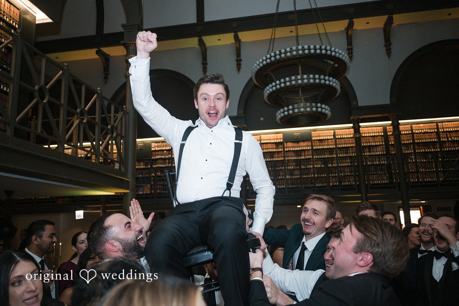 A groom being lifted up in a chair by guests during a wedding reception celebration.